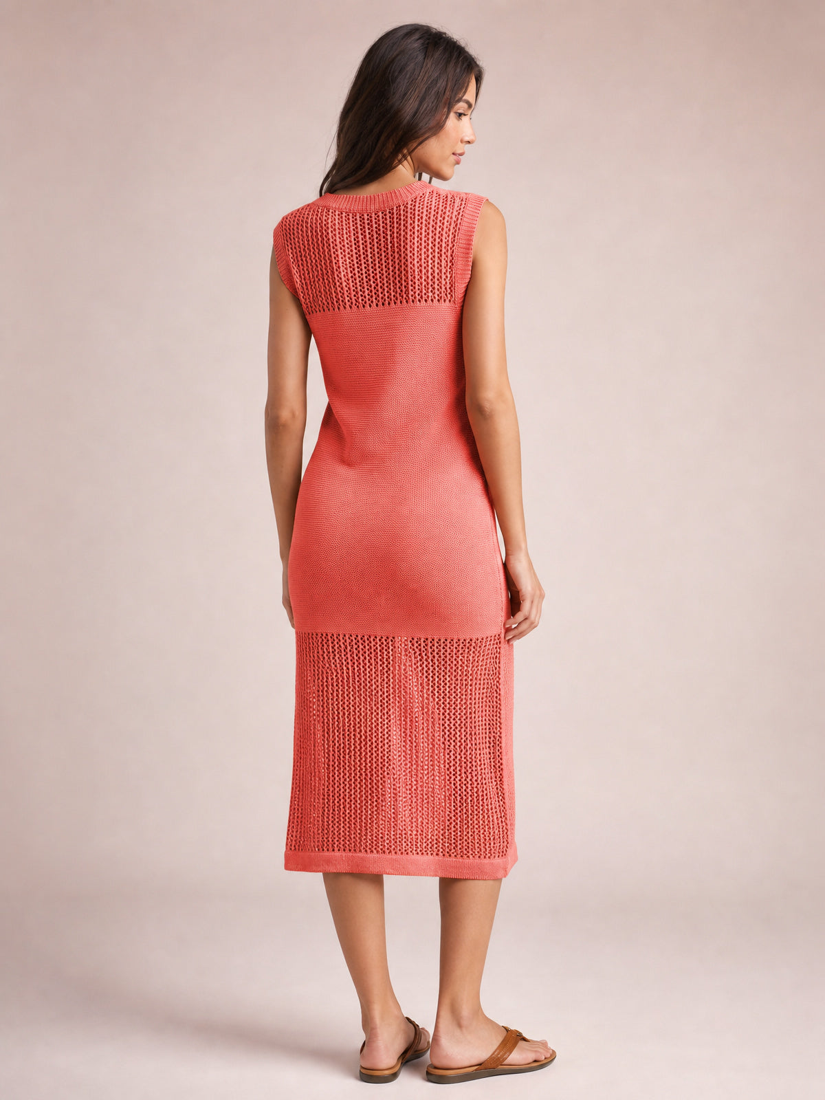 Model poses from the back in a coral sleeveless sweater dress with a round neck, openwork knit panels, fitted silhouette, and below-knee hem.