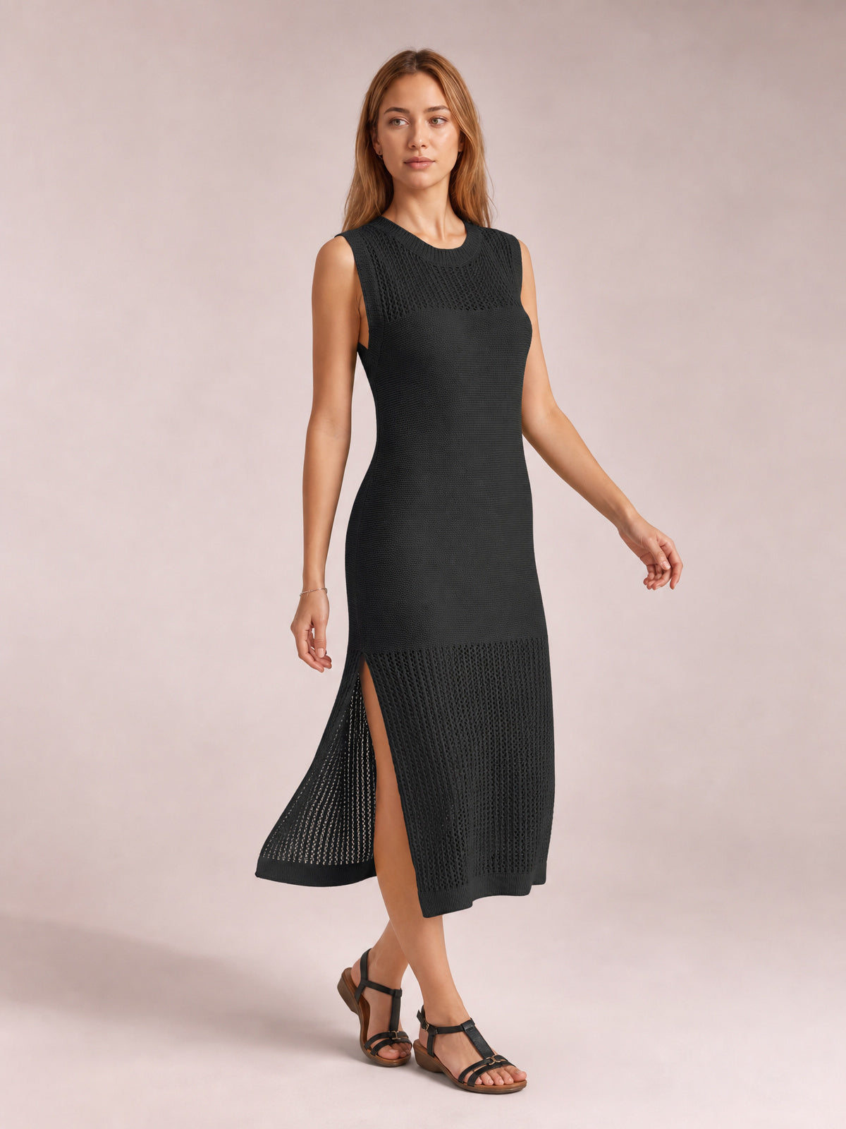 Model poses in a black sleeveless sweater dress with a round neck, fitted midi silhouette, openwork knit panels, and a high side slit.