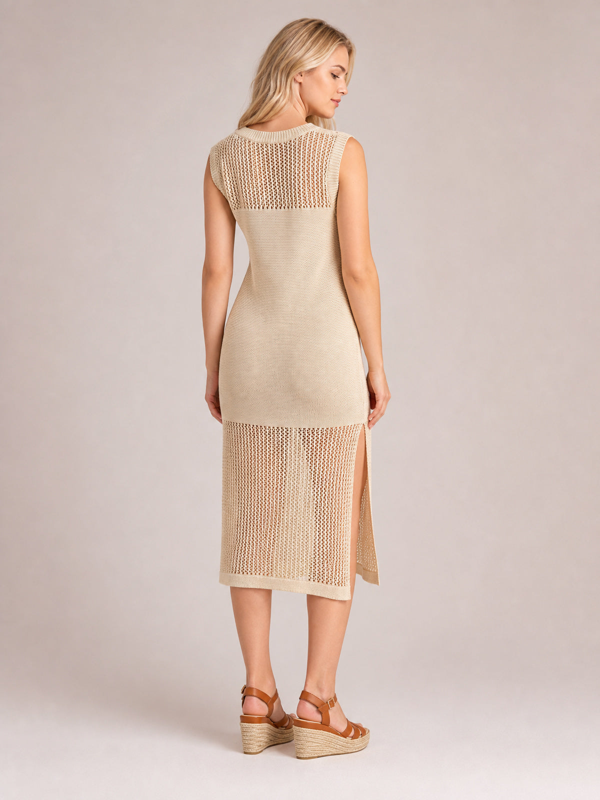 Model poses in a back view wearing a beige sleeveless sweater dress with a round neck, fitted midi silhouette, openwork panels, and side slits.