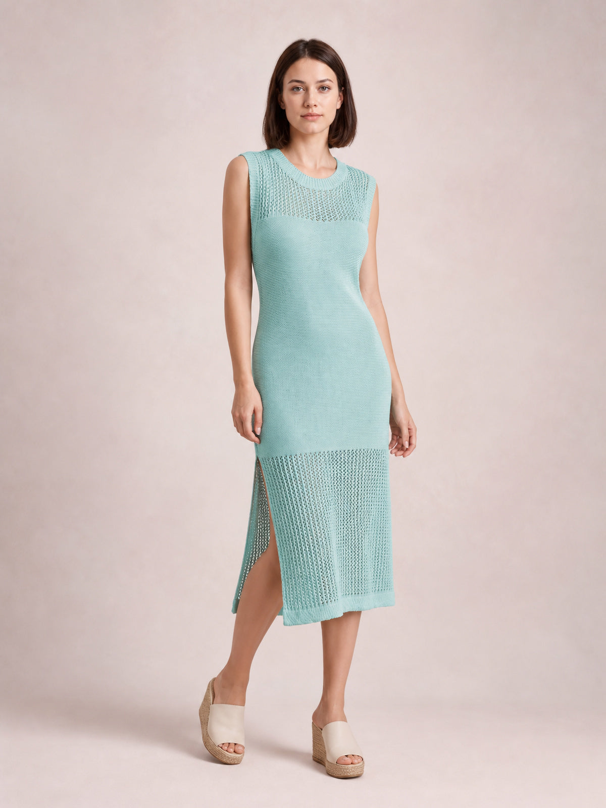 Model poses in a sleeveless aqua sweater dress with a round neckline, fitted midi silhouette, openwork knit panels, and a high side slit.