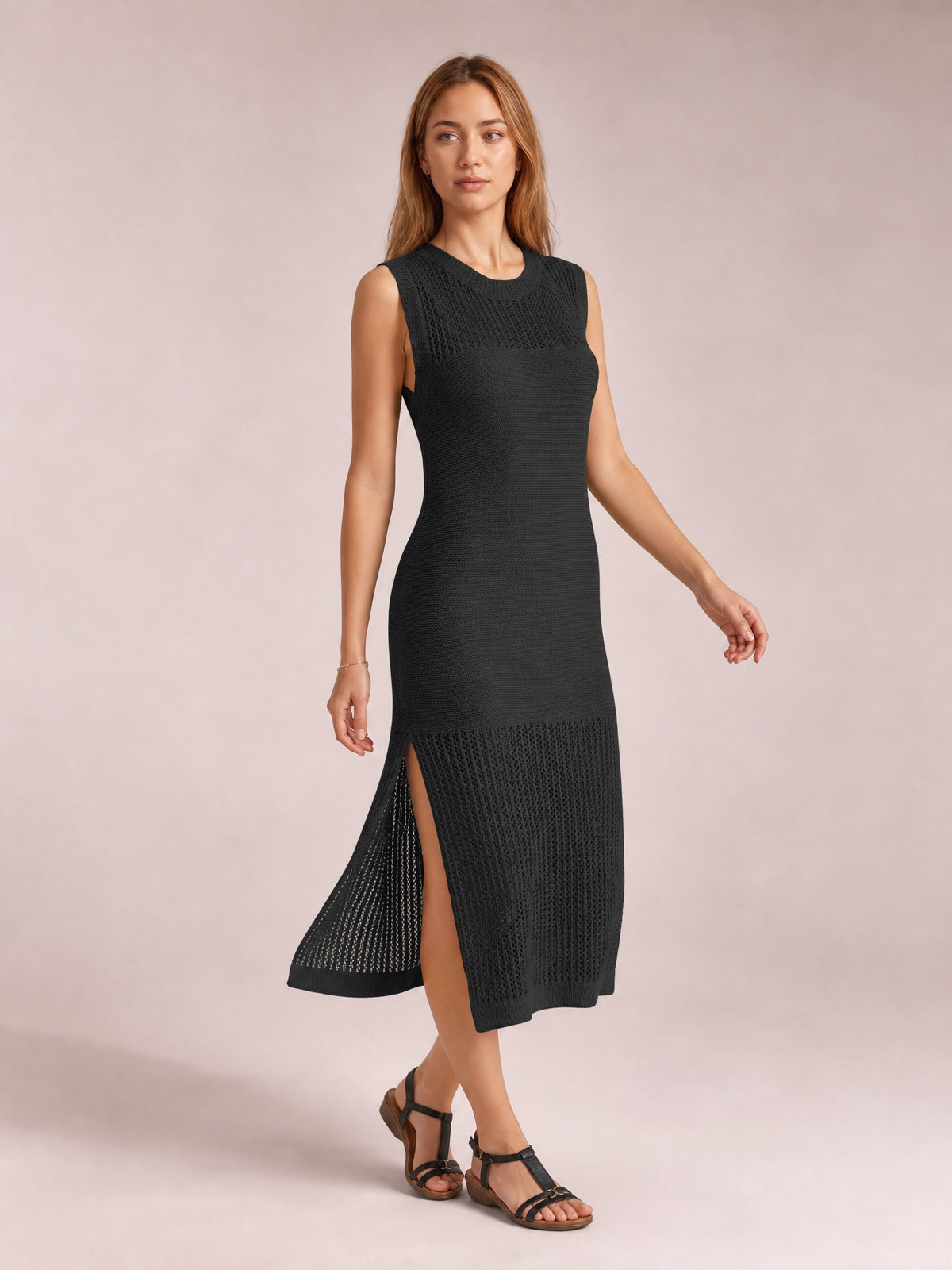 Model poses in a black sleeveless sweater dress with a round neck, fitted midi silhouette, openwork knit panels, and a high side slit.