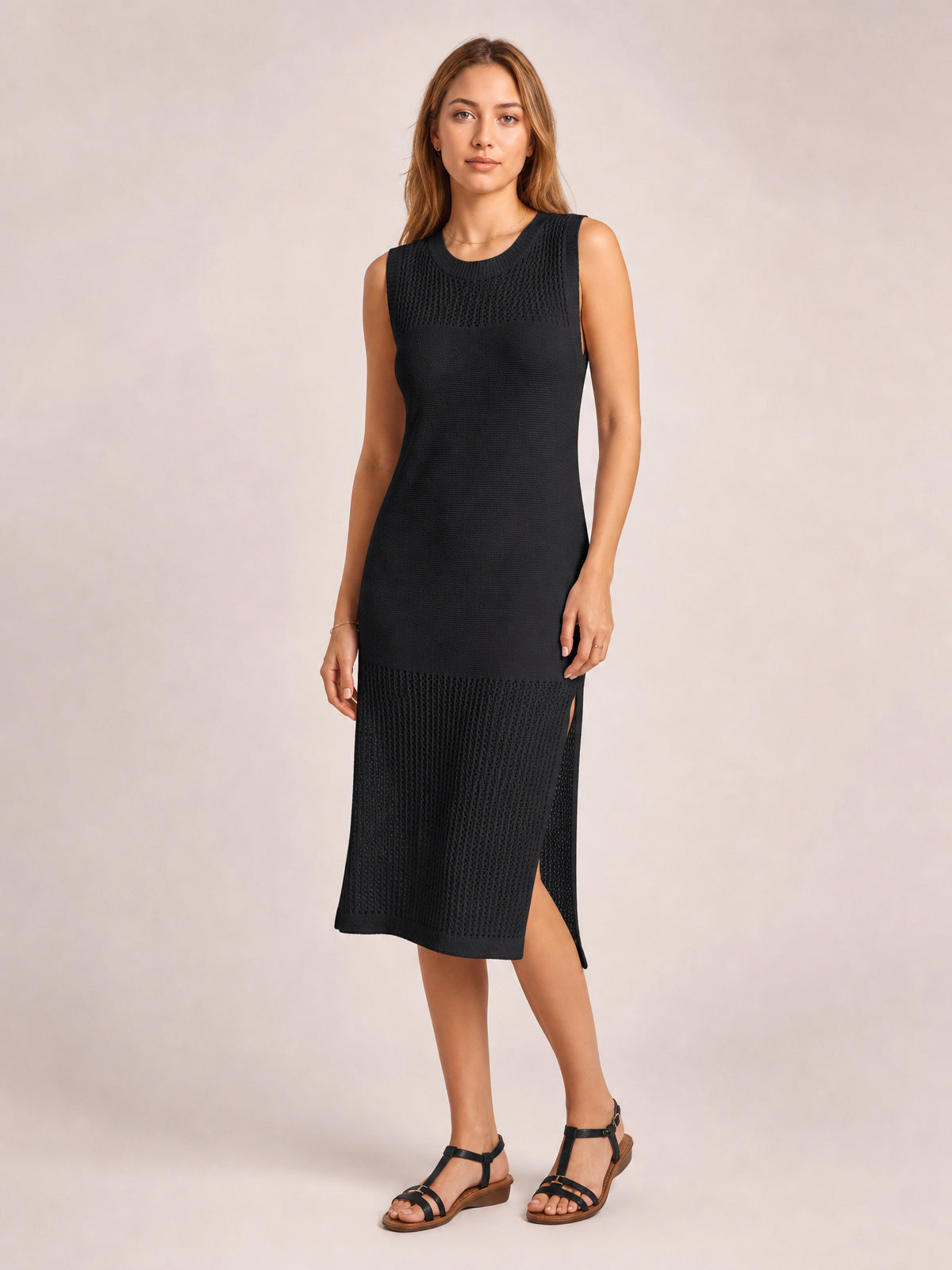 Model in a black sleeveless sweater dress with a round neck, elongated straight silhouette, openwork knit panels, and side slits, standing with one leg crossed.