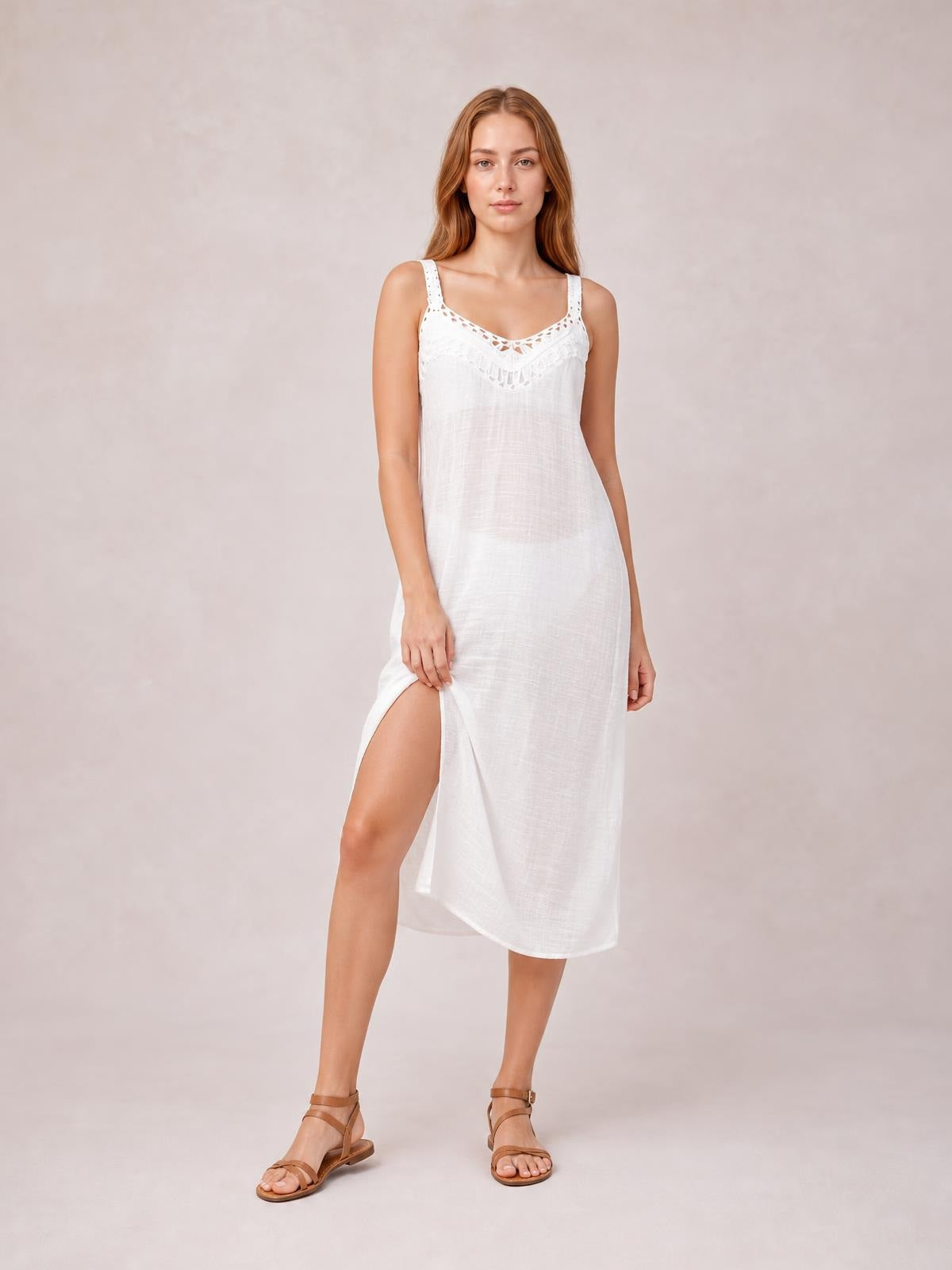 A model stands in a white semi-sheer scoop neck cover up with wide straps, lace neckline detail, straight midi silhouette, and a side slit she holds open.