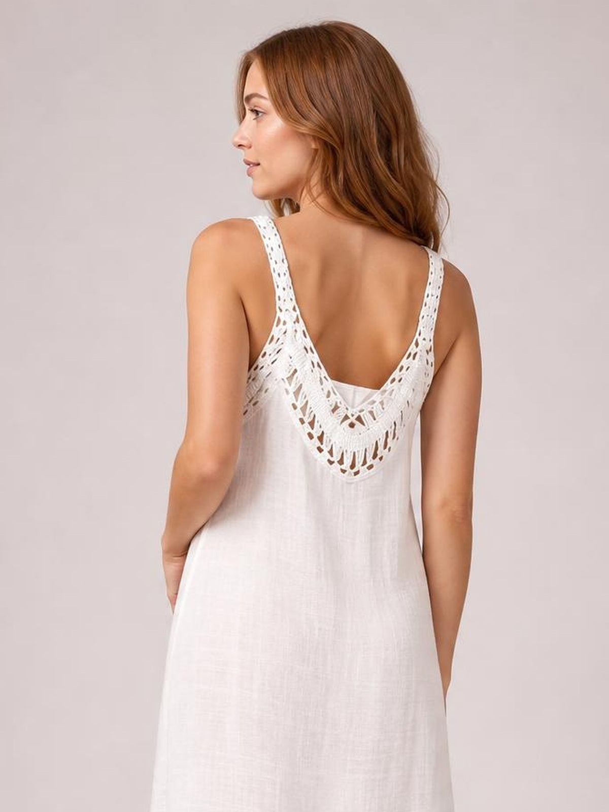 A model faces away in a white semi-sheer scoop neck cover up with wide straps, a flowing loose silhouette, and intricate lace detail across the back.