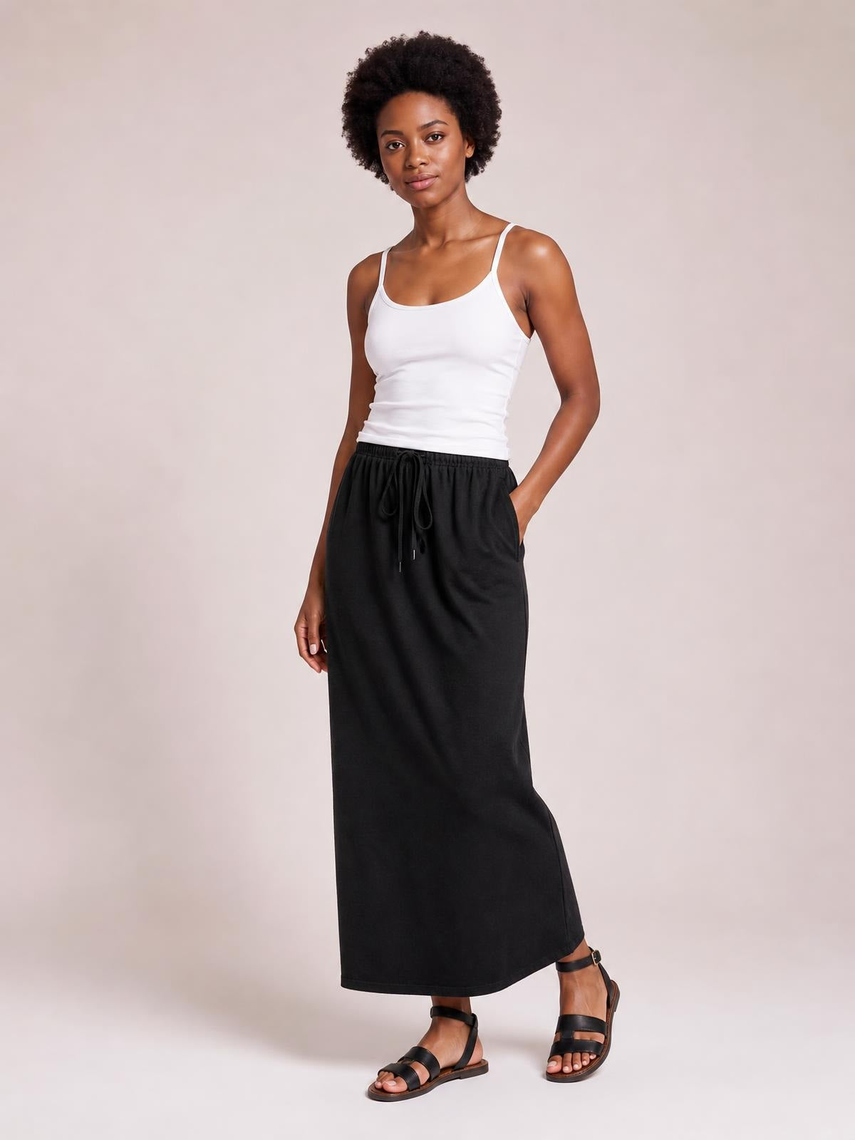 Model wears a black maxi slit skirt with an adjustable drawstring waist, relaxed straight silhouette, and side pocket, posing with one hand tucked in.