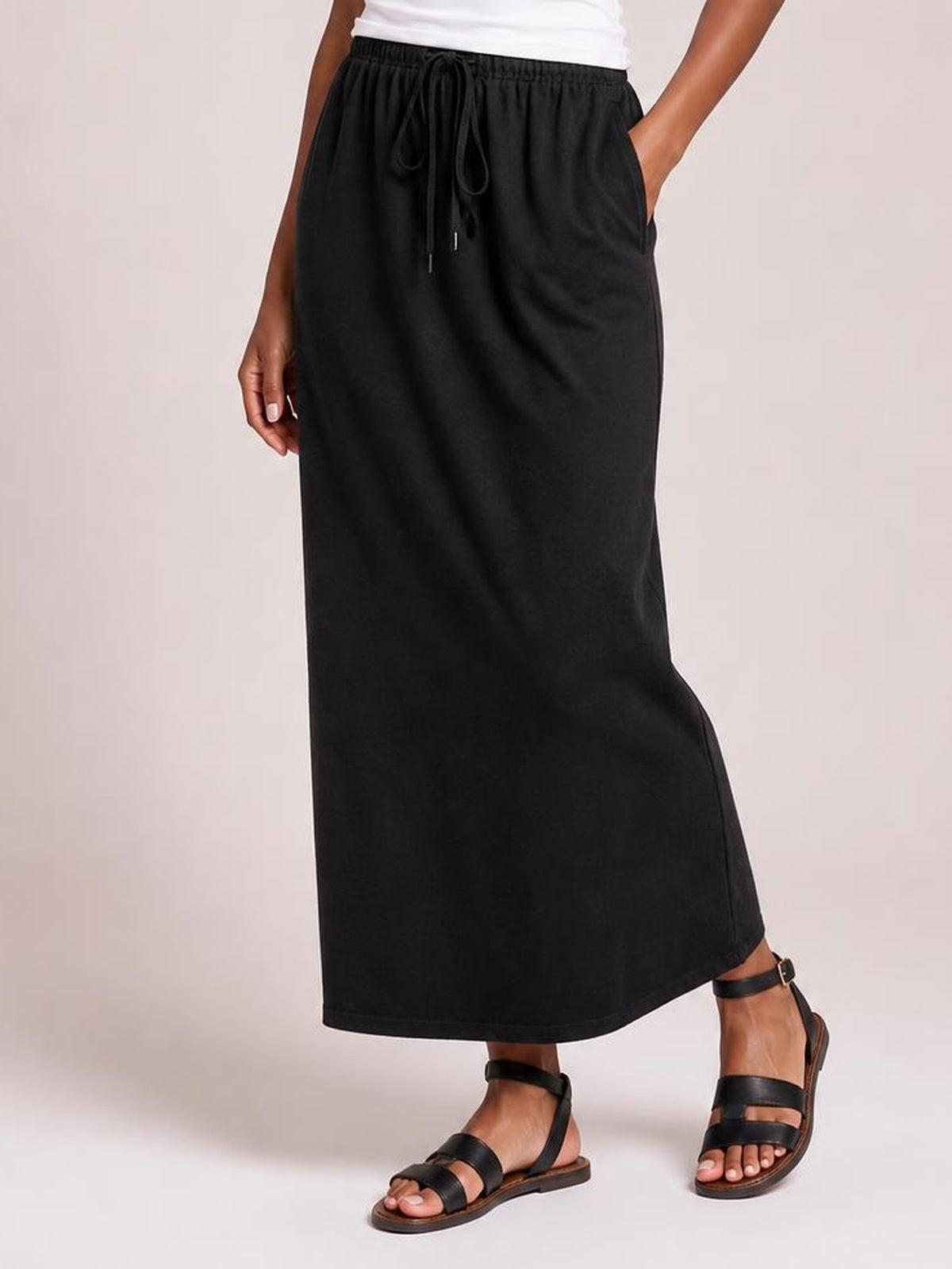 A model wears a black maxi slit skirt with an adjustable drawstring waist, relaxed straight silhouette, side pockets, and a flowing ankle-length hem.