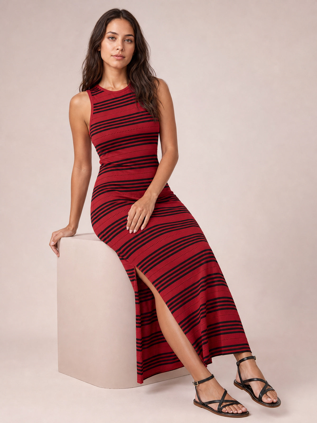 A model sits in a fitted red and black striped midi dress with a round neck, sleeveless cut, streamlined silhouette, and high side slit.