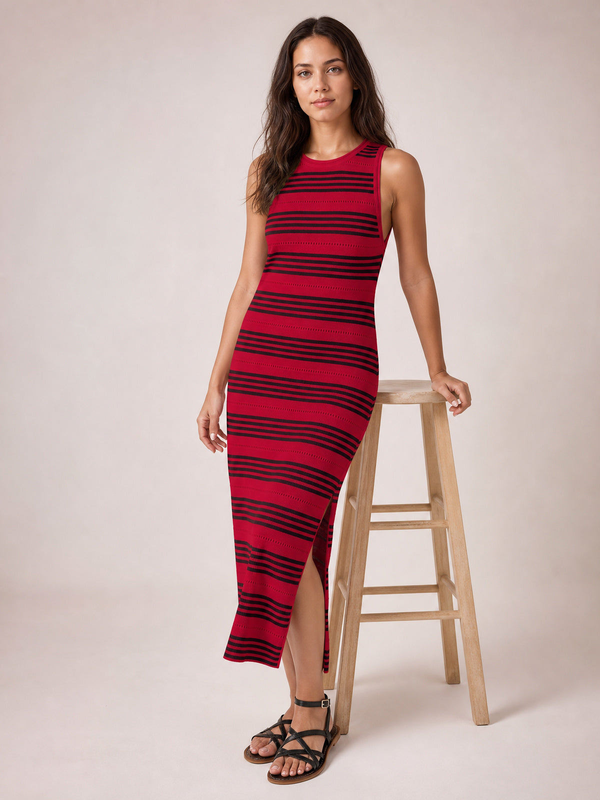 A model leans against a stool in a red and black striped sleeveless midi dress with a round neckline, fitted silhouette, and high side slit.
