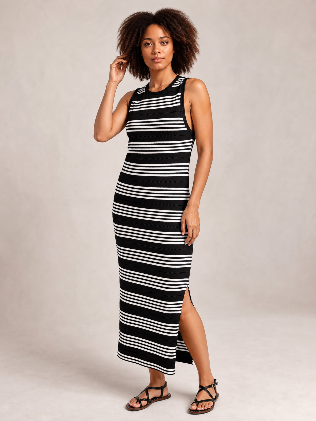 Model poses in a black-and-white striped sleeveless midi dress with a round neck, fitted silhouette, and side slit, touching her hair.