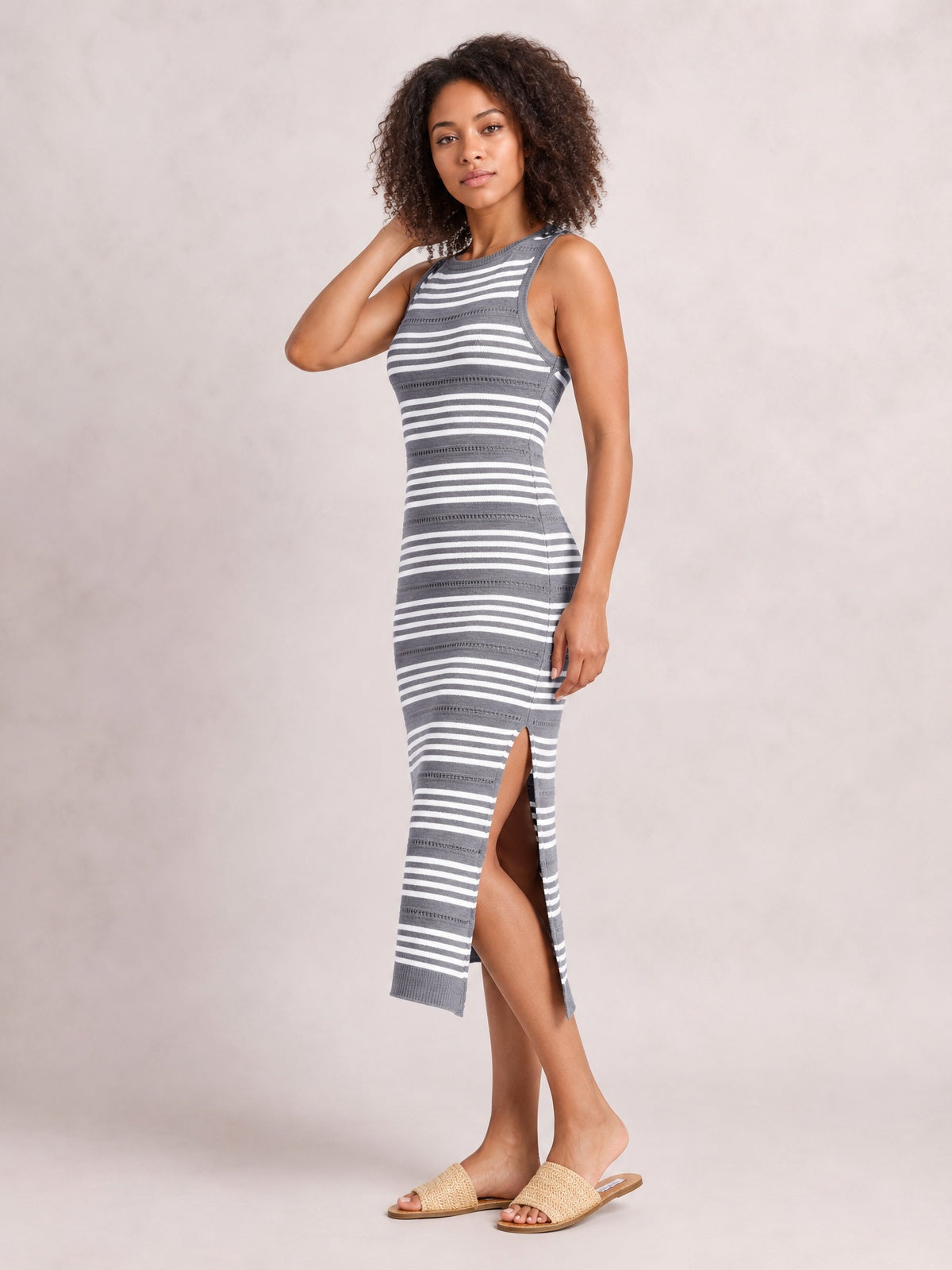 Model poses in a gray and white striped sleeveless midi dress with a round neck, fitted silhouette, and high side slit.