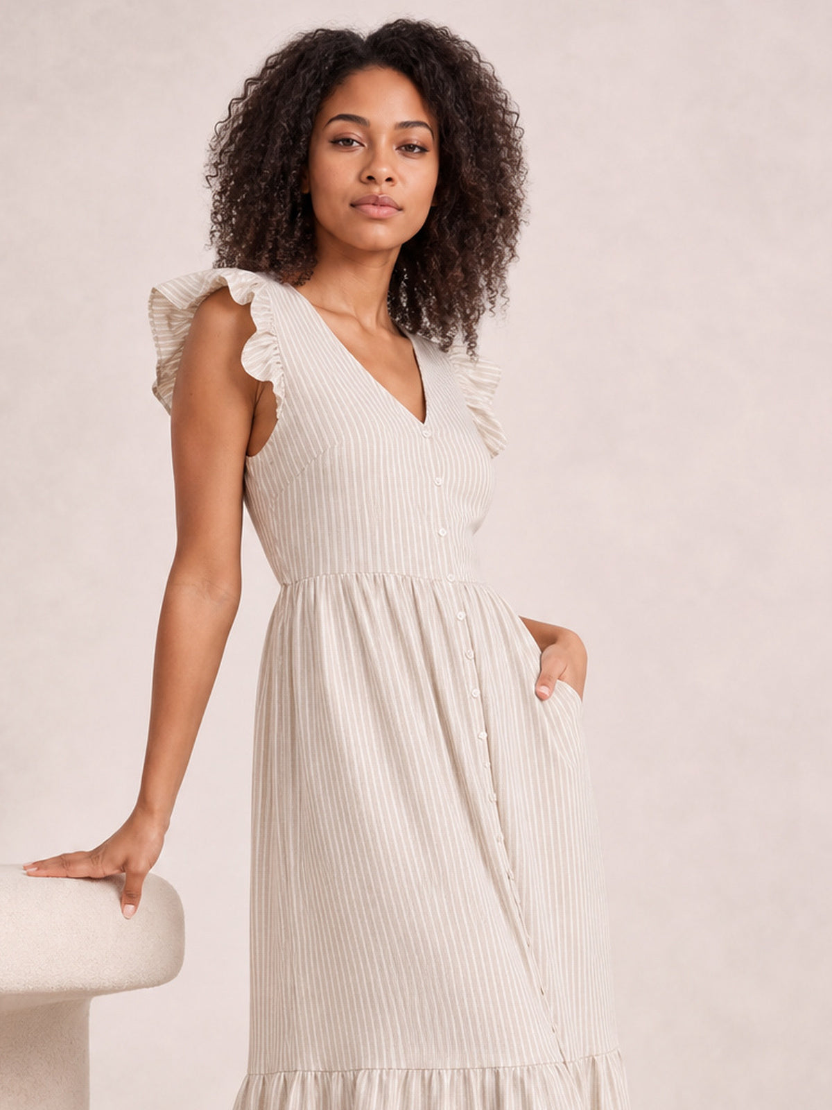 A model poses in a light beige striped V-neck midi dress with ruffled cap sleeves, button-front construction, a defined waist, tiered skirt, and side pockets.