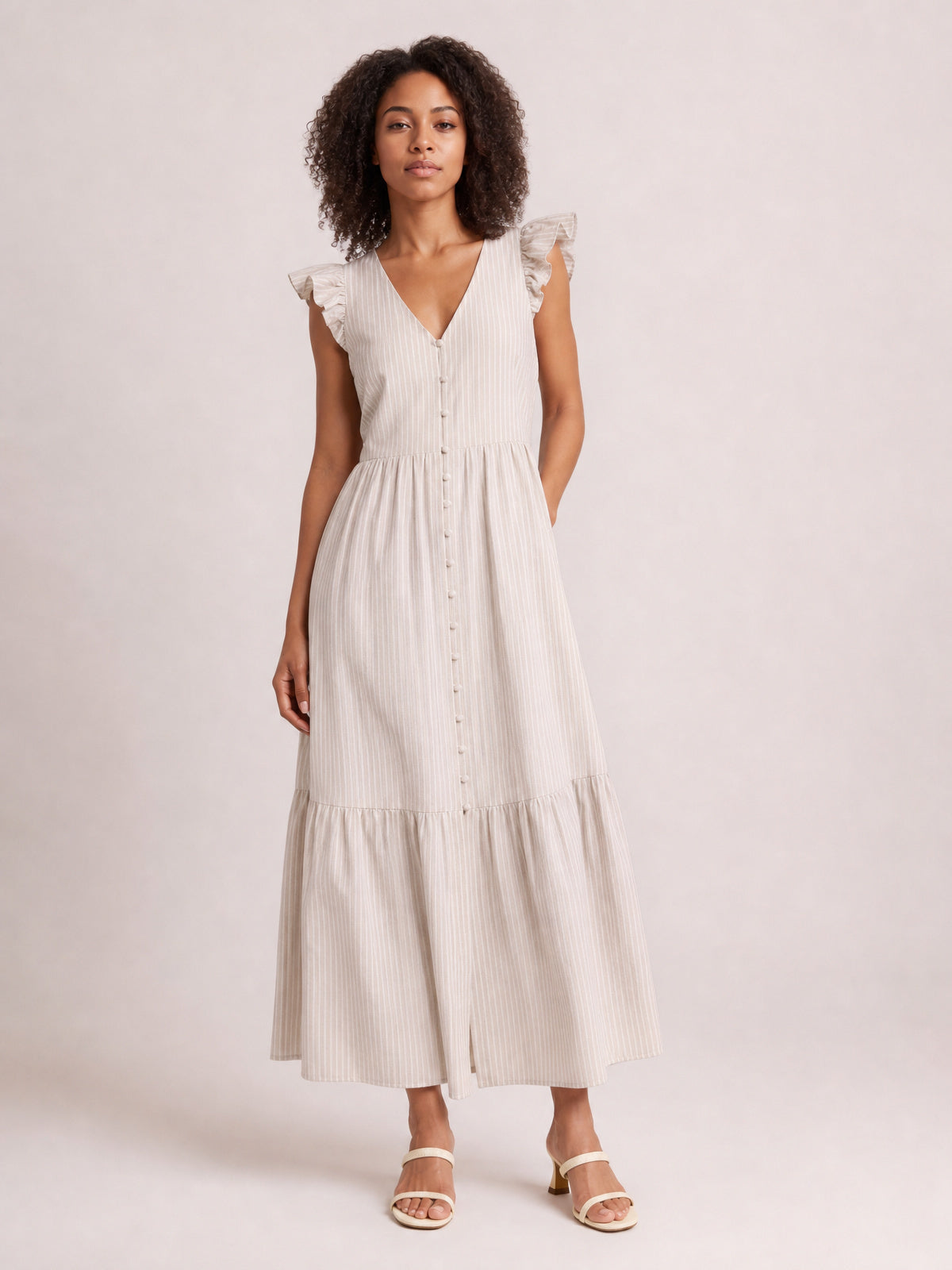 Model poses in a light beige striped V-neck midi dress with ruffled cap sleeves, buttoned bodice, defined waist, tiered skirt, side pockets, and front slit.