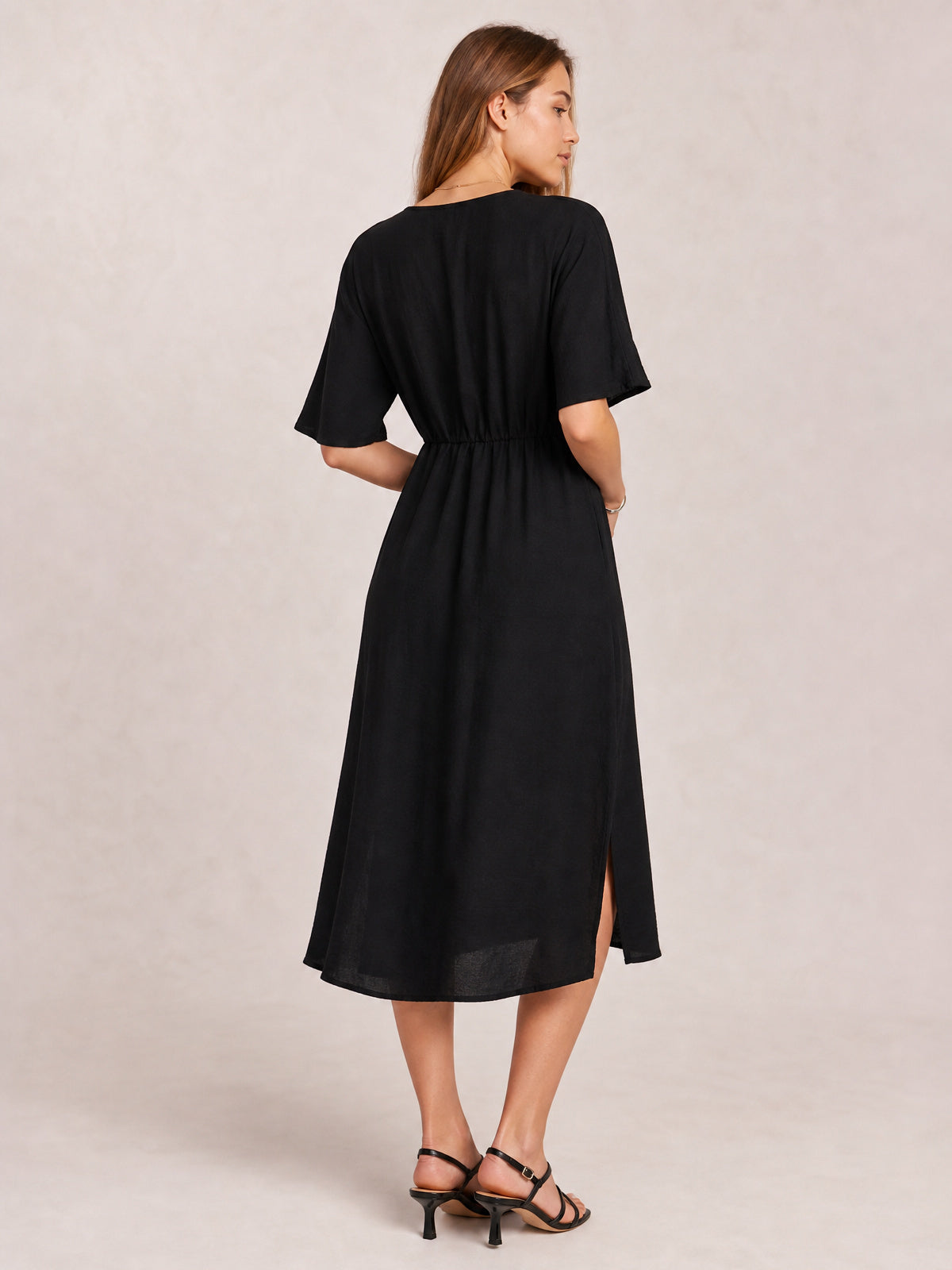 A model looks over her shoulder in a black half-sleeve midi dress with a cinched waist, flowing skirt, and visible side slit.