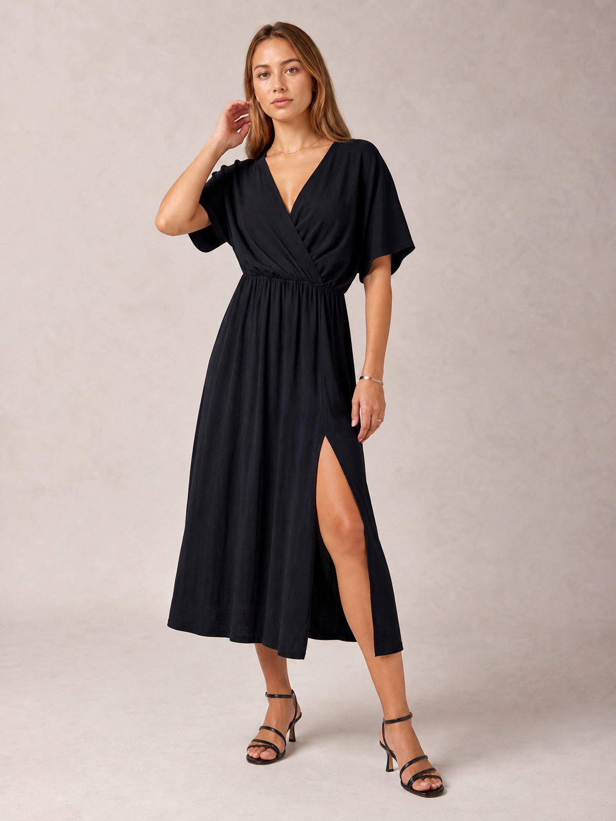 Model poses in a black surplice midi dress with a wrap-inspired V-neckline, gathered waist, flowing half sleeves, and a high side slit.