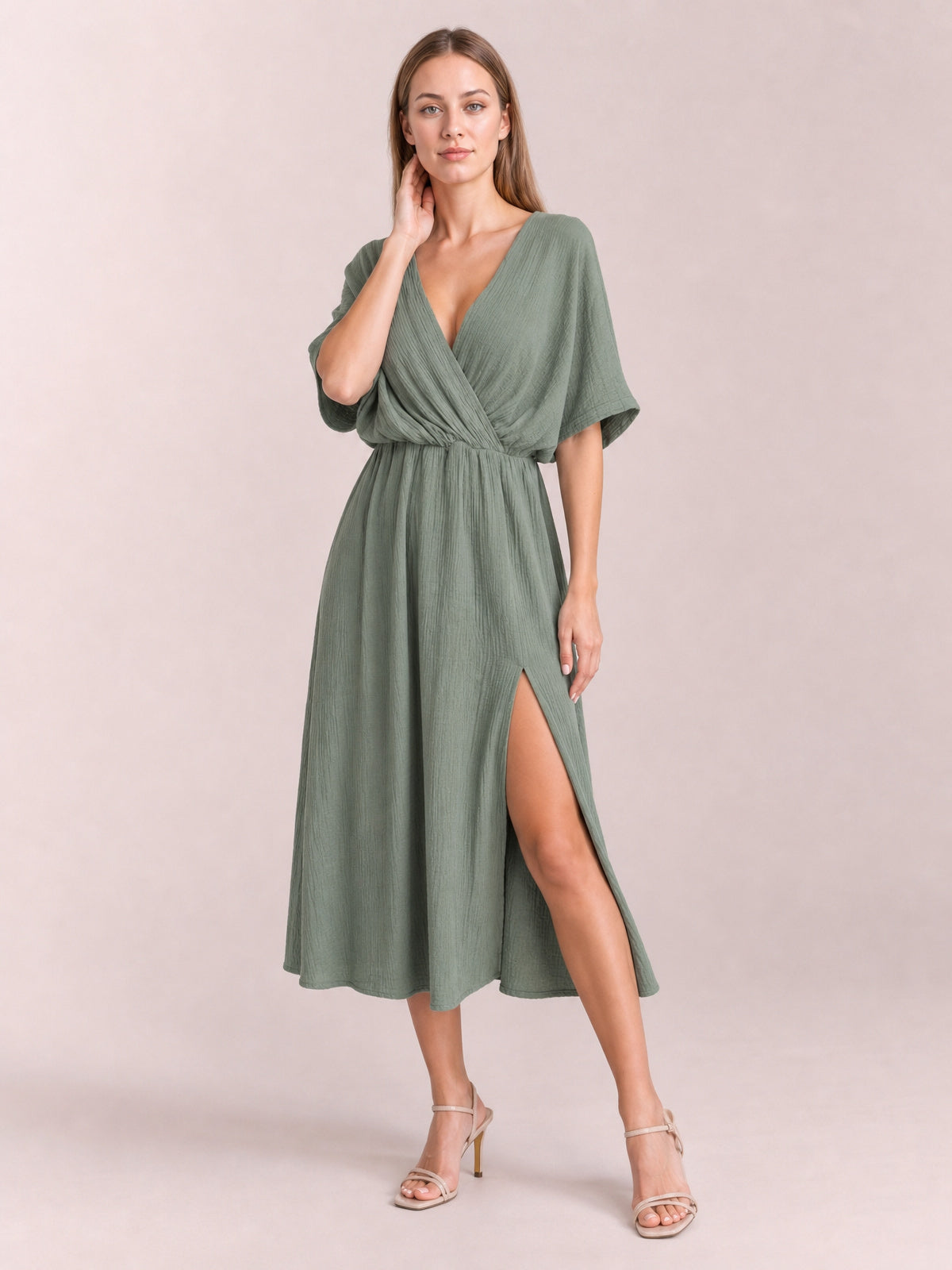 Model poses in a sage green slit surplice midi dress with a wrap-inspired V-neck, gathered waist, relaxed half sleeves, and flowing skirt.