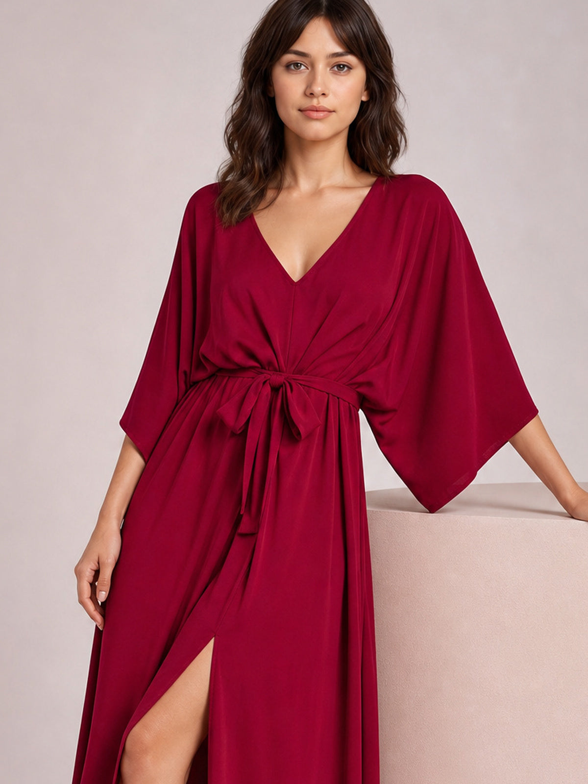 A model poses in a burgundy V-neck dress with a tied gathered waist, flowing silhouette, wide three-quarter sleeves, and a high side slit, resting one hand on a ledge.