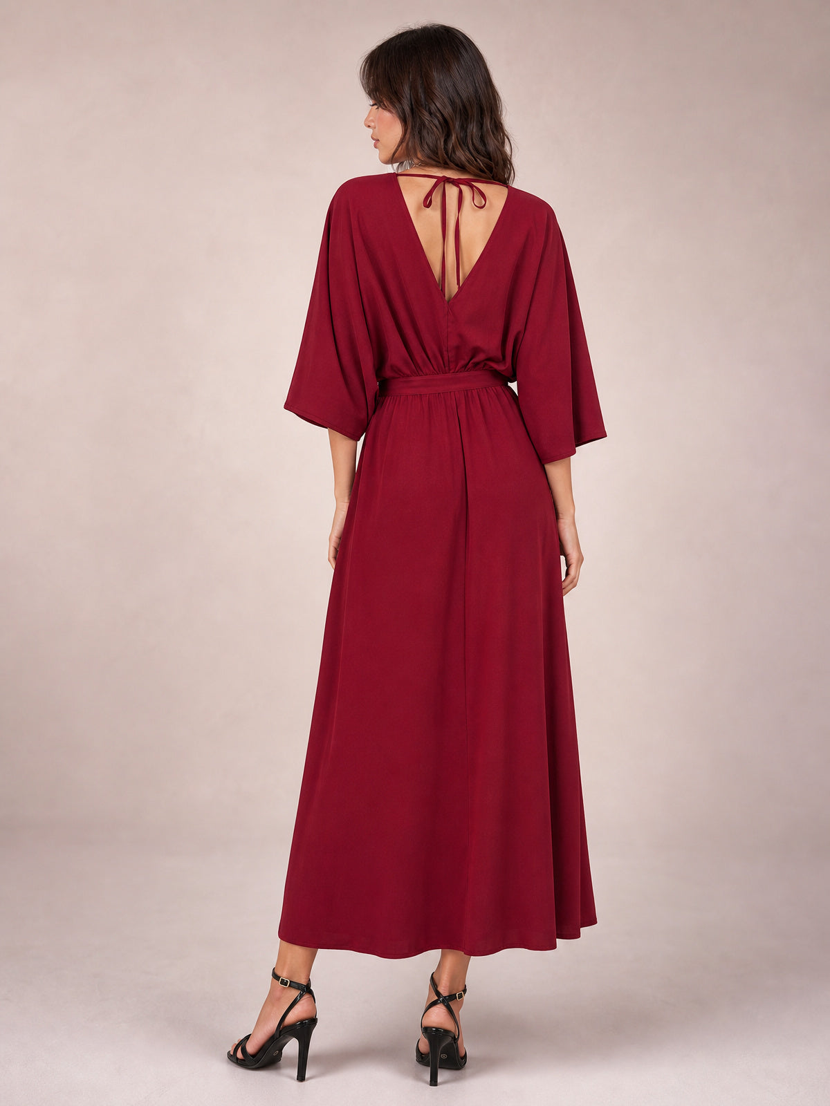 Model viewed from behind in a burgundy V-neck dress with three-quarter sleeves, a defined tied waist, flowing maxi skirt, and open back tie detail.
