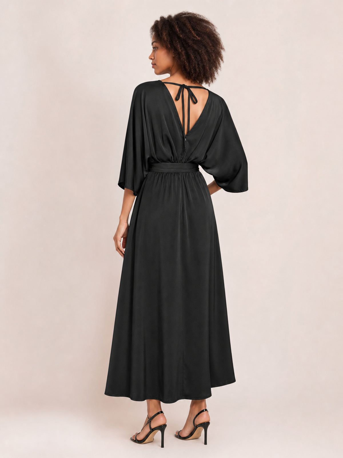 A model poses in a black V-neck dress with a tied open back, softly gathered waist, flowing maxi silhouette, and relaxed three-quarter sleeves.