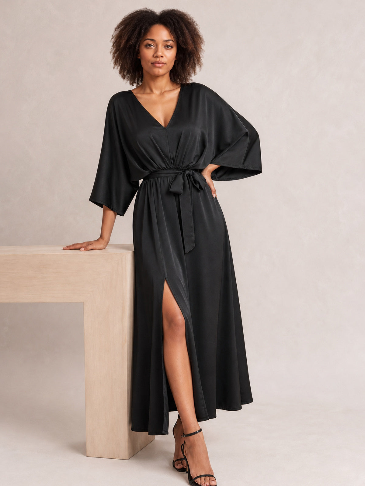 Model in a black slit tied V-neck dress with flowing maxi silhouette, softly gathered waist, bow belt, and relaxed three-quarter sleeves, posing beside a pedestal.