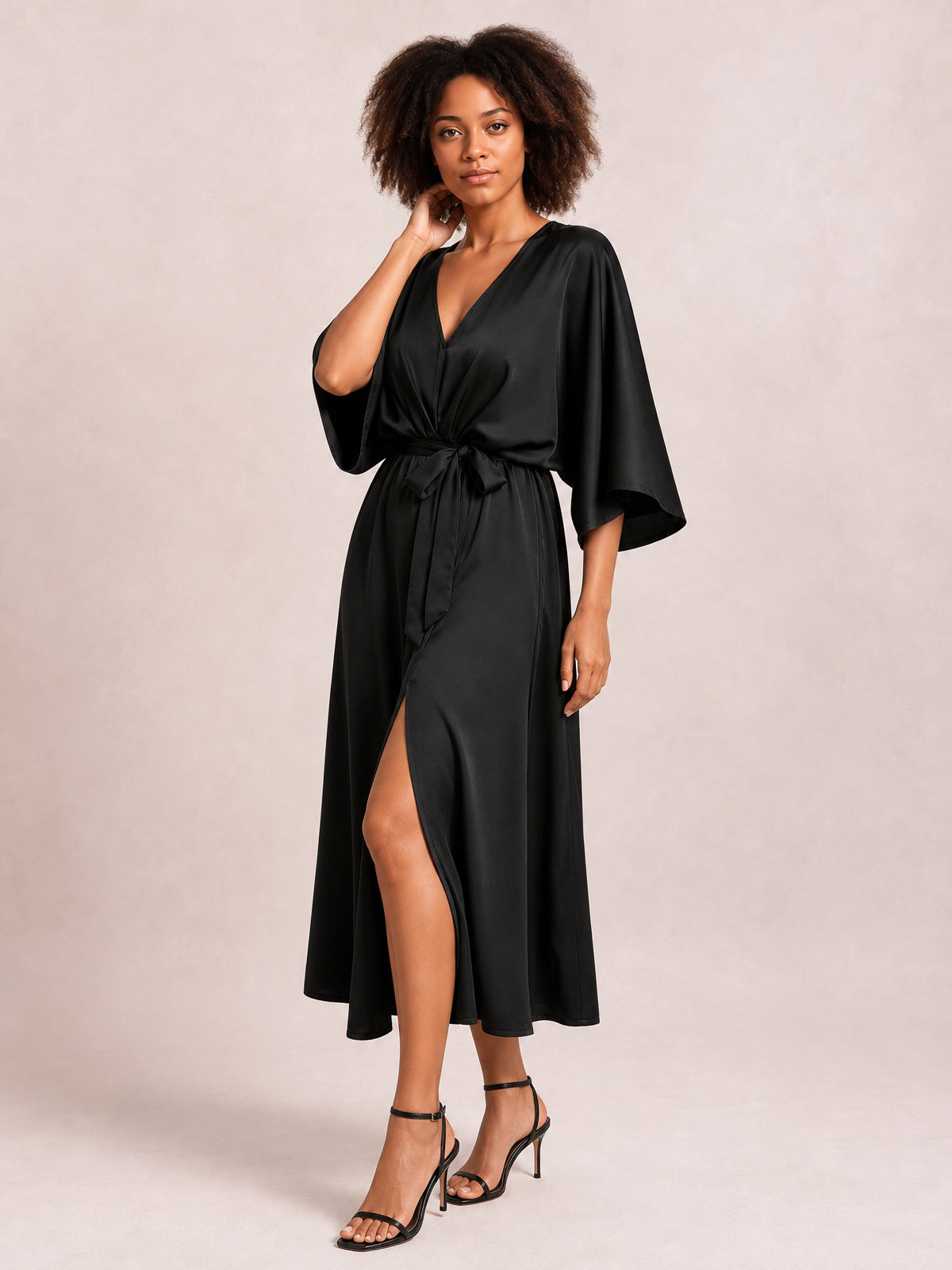 Model poses in a black V-neck dress with flowing three-quarter sleeves, a tied waist, flattering gathered silhouette, and daring side slit.