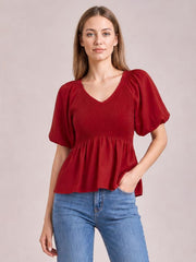 A model wears a red V-neck peplum blouse with a smocked bodice, short balloon puff sleeves, and a softly flared hem.
