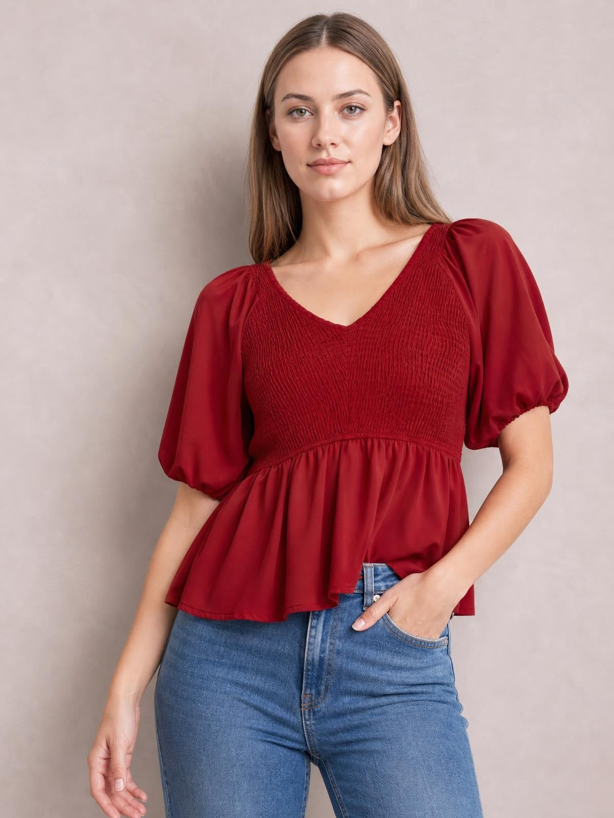 Model wearing a burgundy smocked V-neck peplum blouse with short puffed balloon sleeves and a flared waist, posing with one hand in her jeans pocket.