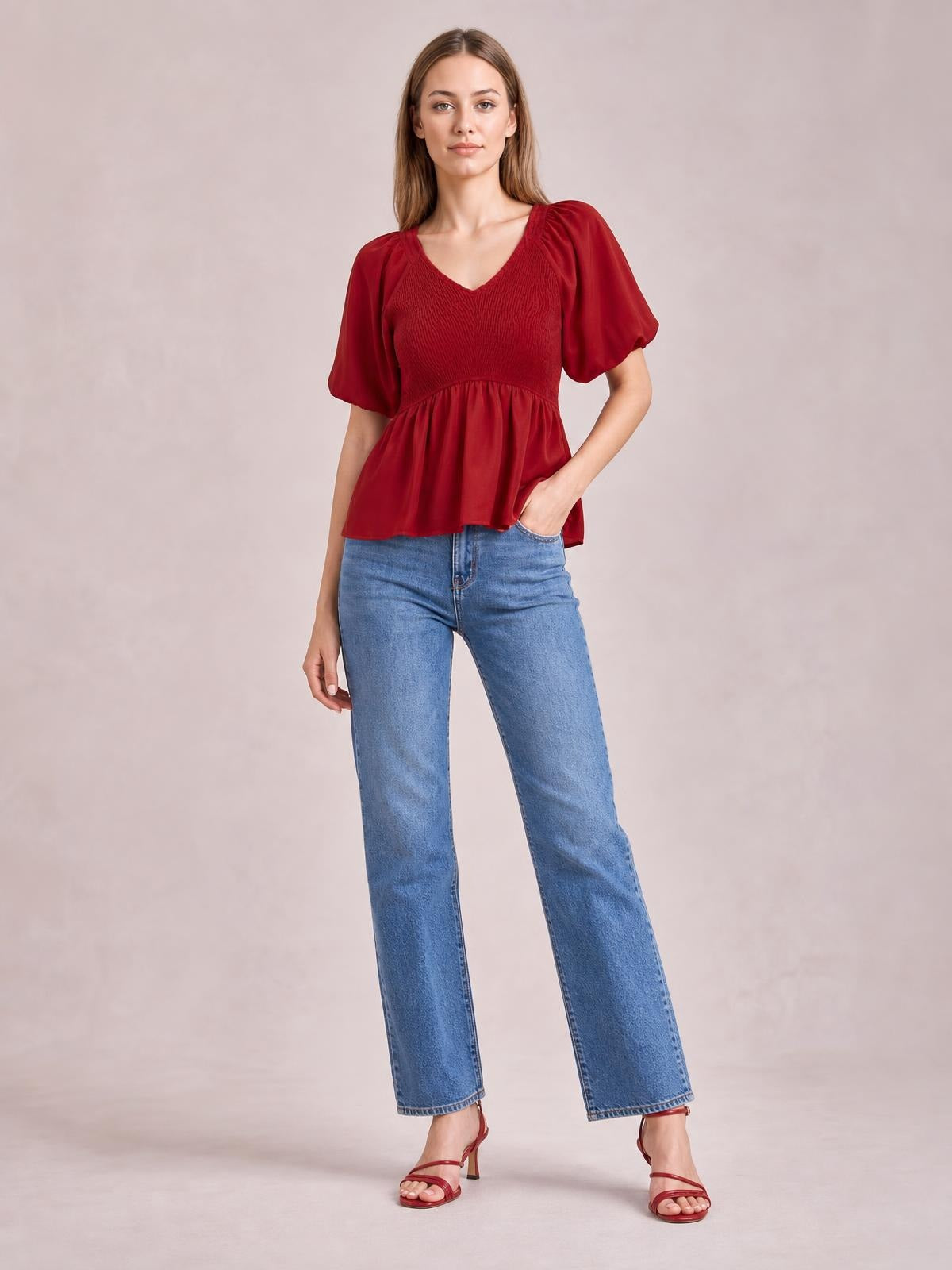 A model poses in a deep red smocked peplum blouse with a V-neck and short balloon puff sleeves, paired with jeans and one hand in a pocket.