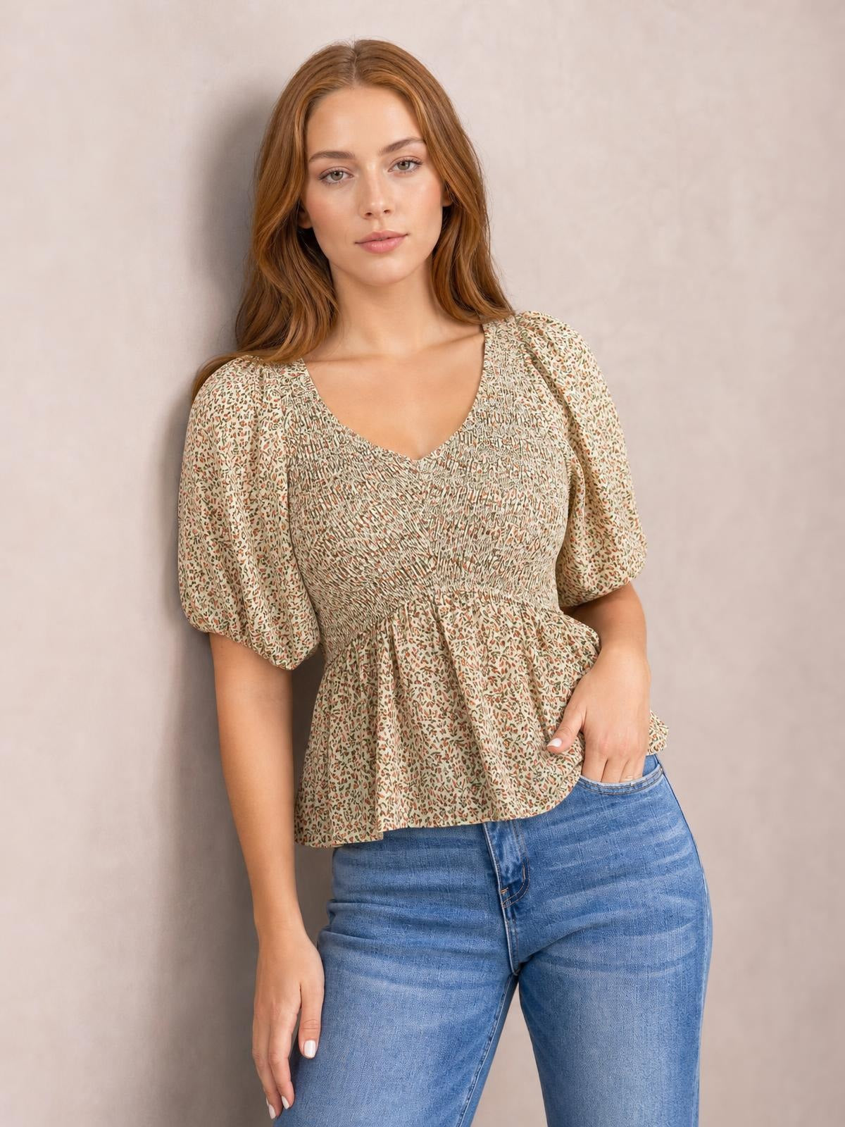 A model stands with one hand in her jeans pocket wearing a beige floral peplum blouse with a V-neck, smocked bodice, and short balloon puff sleeves.