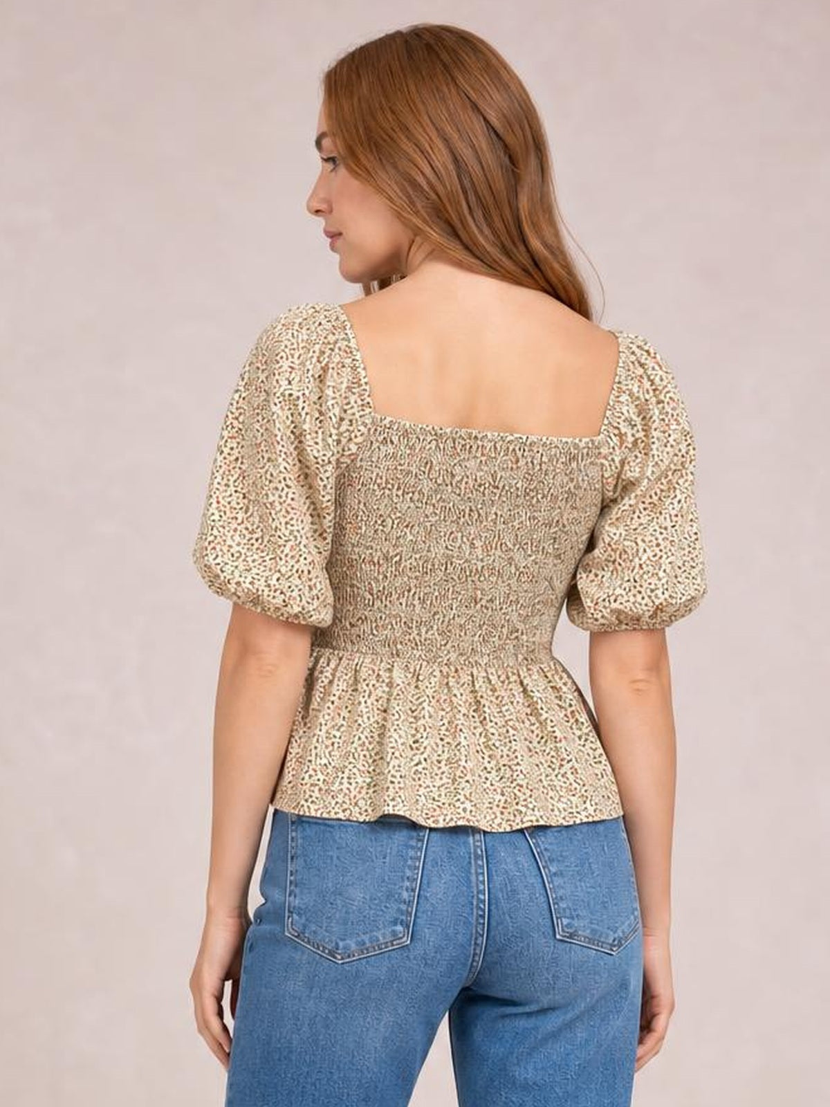 Model shown from behind wears a beige floral smocked peplum blouse with short puffed balloon sleeves, a fitted bodice, and flared hem.