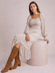 Model seated in a floral midi dress with a square neck, smocked bodice, long puff sleeves, gathered waist, flowing skirt, and flounced hem.