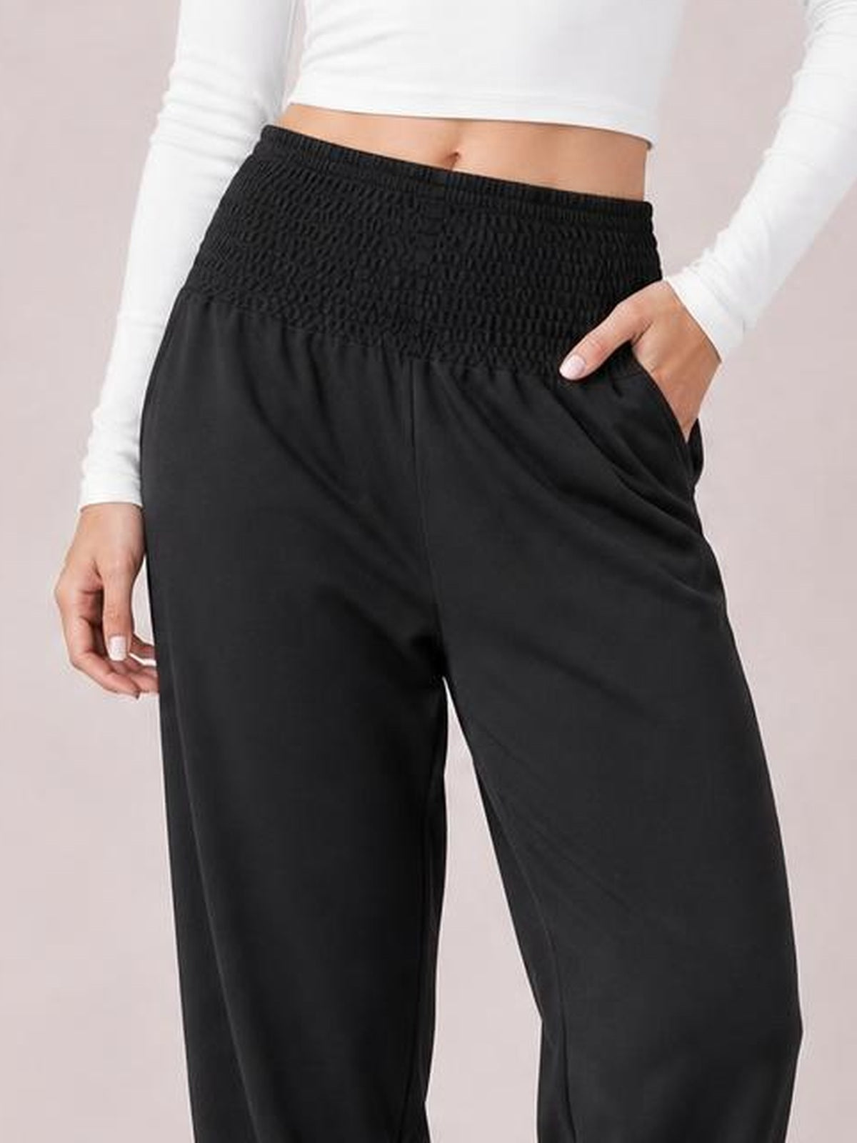 A model wears black smocked high rise joggers with a gathered waistband, loose straight-leg silhouette, and side pockets, resting one hand inside.