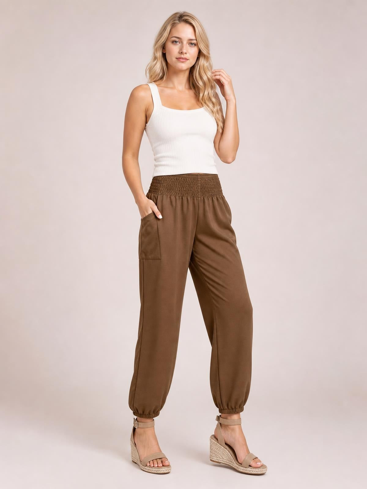 Model wears brown smocked high rise joggers with a relaxed tapered silhouette, side pockets, elastic cuffs, and one hand tucked into a pocket.