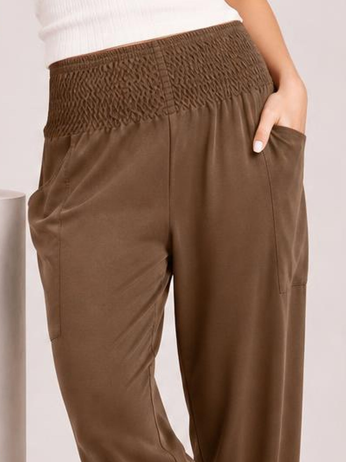 Model wearing brown high-rise joggers with a smocked waistband, relaxed straight-leg silhouette, side pockets, and visible seam construction.