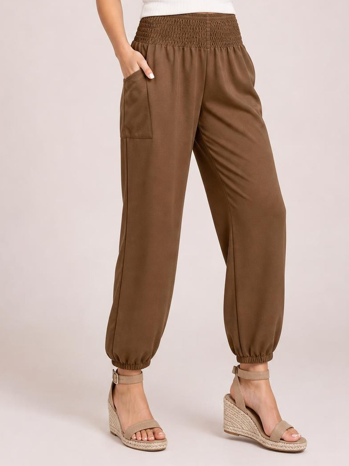 Model wears brown high-rise joggers with a smocked waistband, side pockets, relaxed legs, and elasticized ankle cuffs, standing with one hand in a pocket.