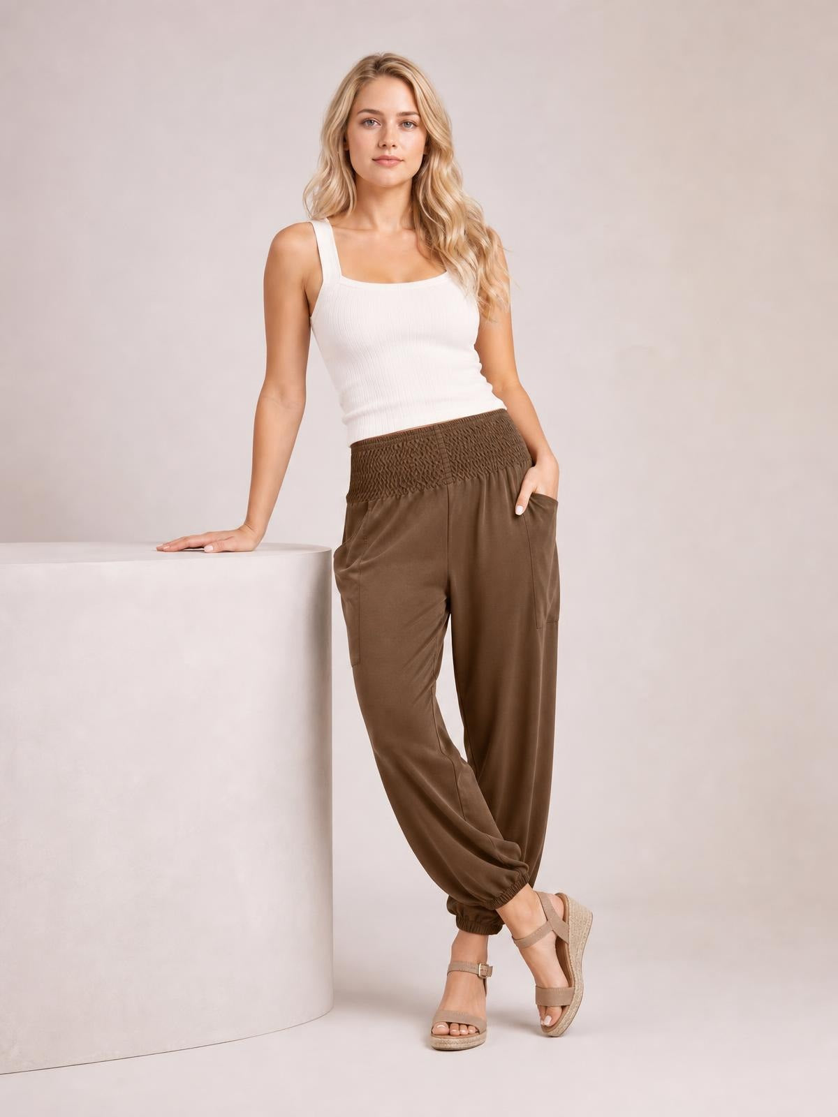 Model poses in brown smocked high-rise joggers with side pockets, a relaxed straight-leg silhouette, and elasticized cuffs, paired with a white tank top.