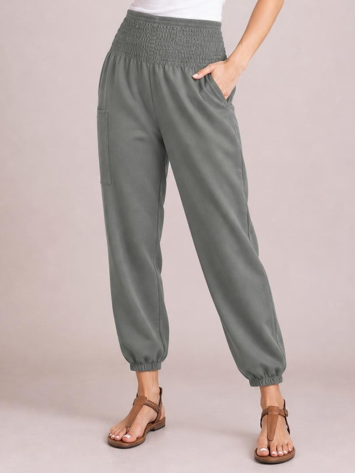A model wears gray high-rise smocked joggers with a relaxed tapered silhouette, elastic cuffs, and practical pockets, posing with one hand tucked inside.