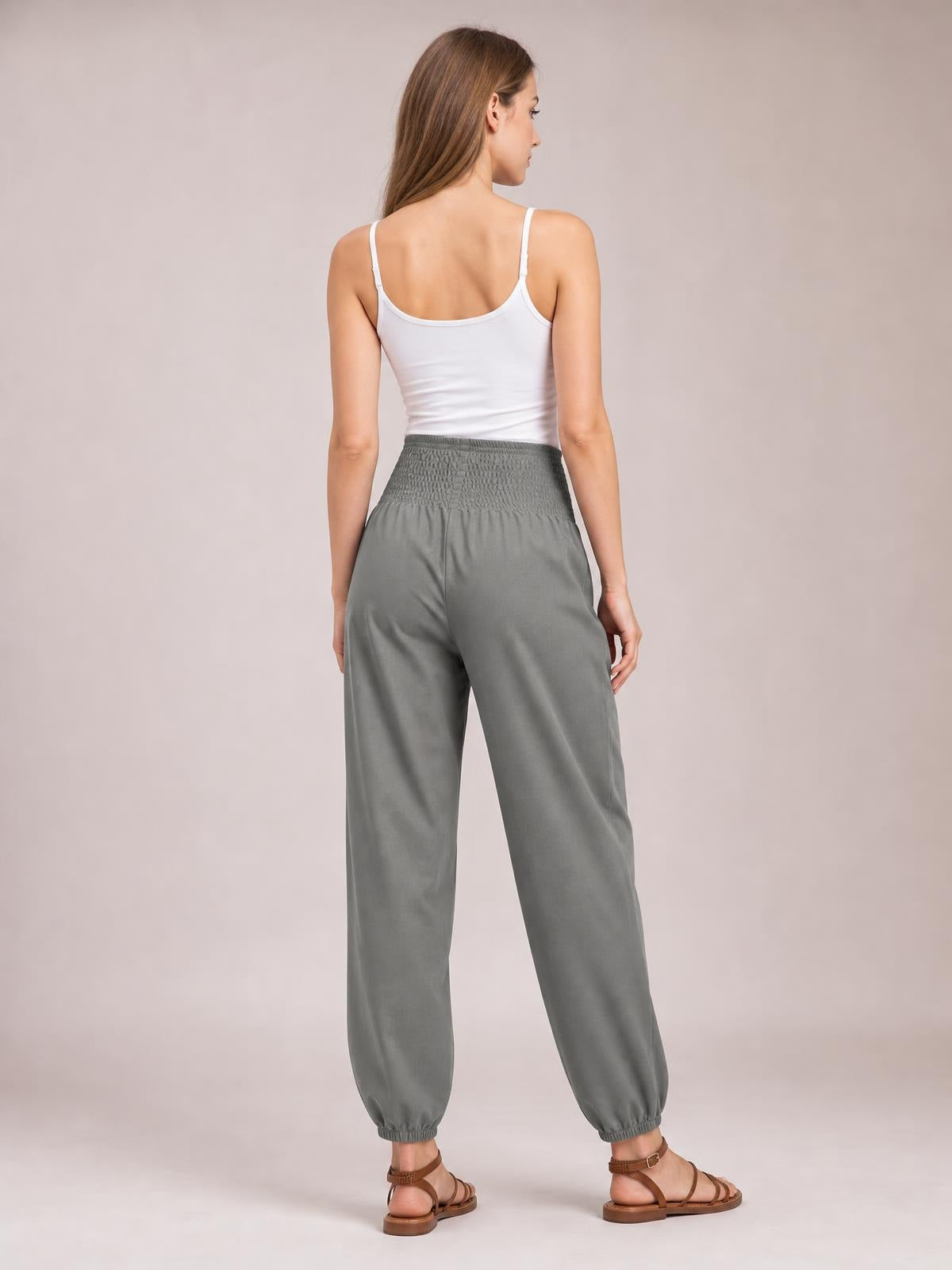 A model stands turned away wearing gray high-rise joggers with a wide smocked waistband, relaxed straight legs, and elasticized ankle cuffs.