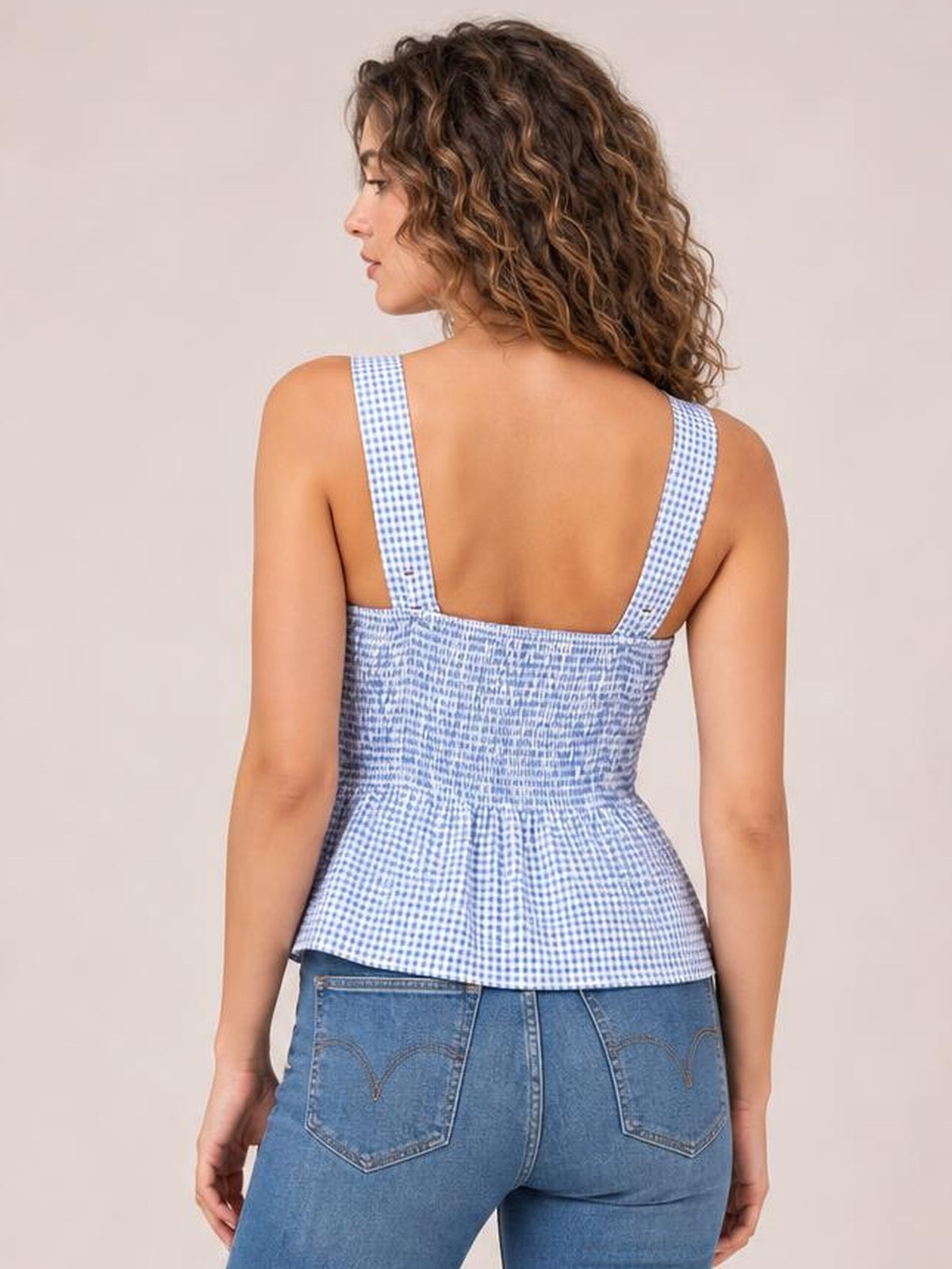 A model faces away wearing a blue-and-white plaid smocked tank with wide shoulder straps, a fitted bodice, and a softly flared hem.