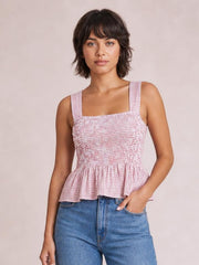 Model wears a pink plaid smocked tank with a square neckline, wide straps, stretchy fitted bodice, and flared peplum hem, posing with one hand in her jeans pocket.