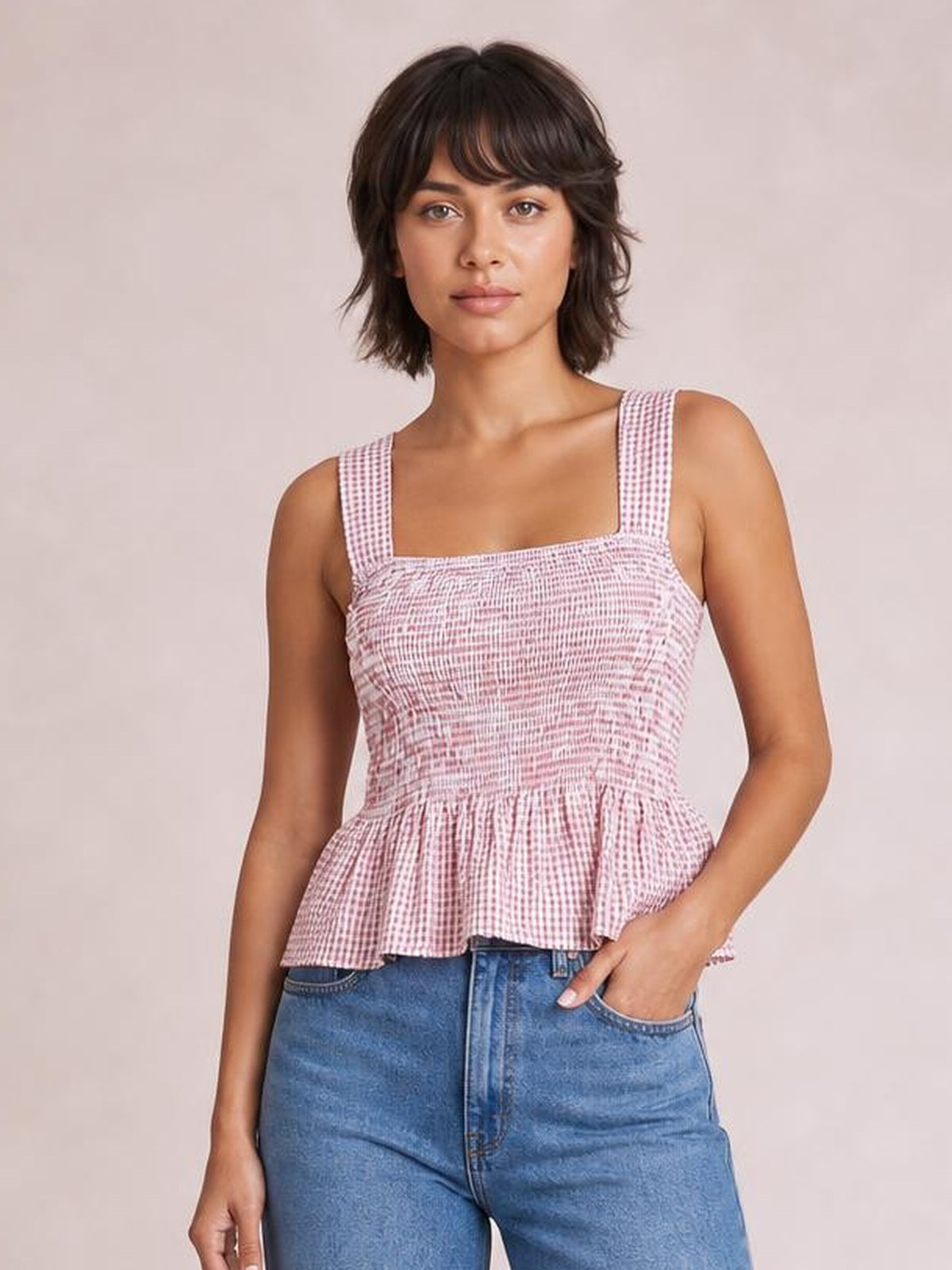 Model wears a pink plaid smocked tank with a square neckline, wide straps, stretchy fitted bodice, and flared peplum hem, posing with one hand in her jeans pocket.