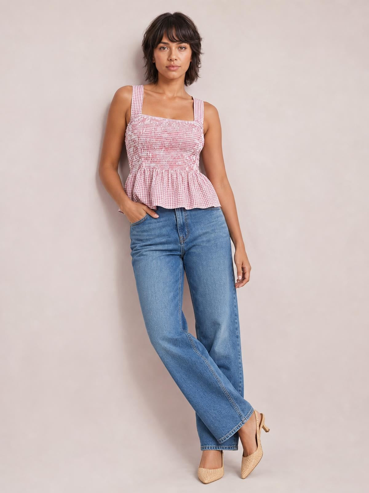 Model poses in a pink and white smocked plaid tank with a square neckline, wide straps, fitted bodice, and flared peplum hem.
