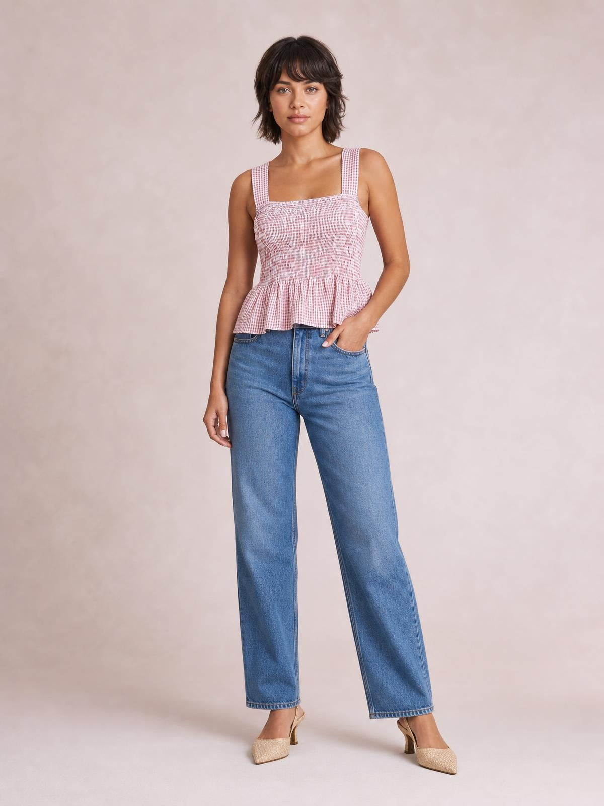 A model wears a pink-and-white plaid smocked tank with a square neckline, wide straps, stretchy fitted bodice, and flared peplum hem while standing with one hand in her pocket.