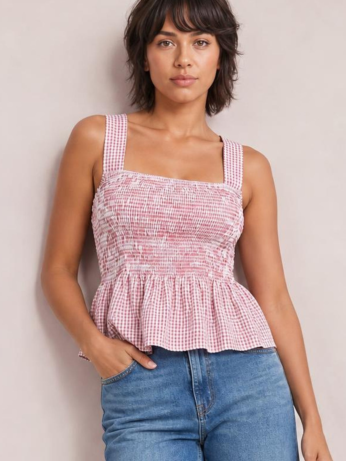 Model wearing a pink-and-white plaid smocked square neck tank with wide straps, a fitted bodice, and a flared peplum hem.