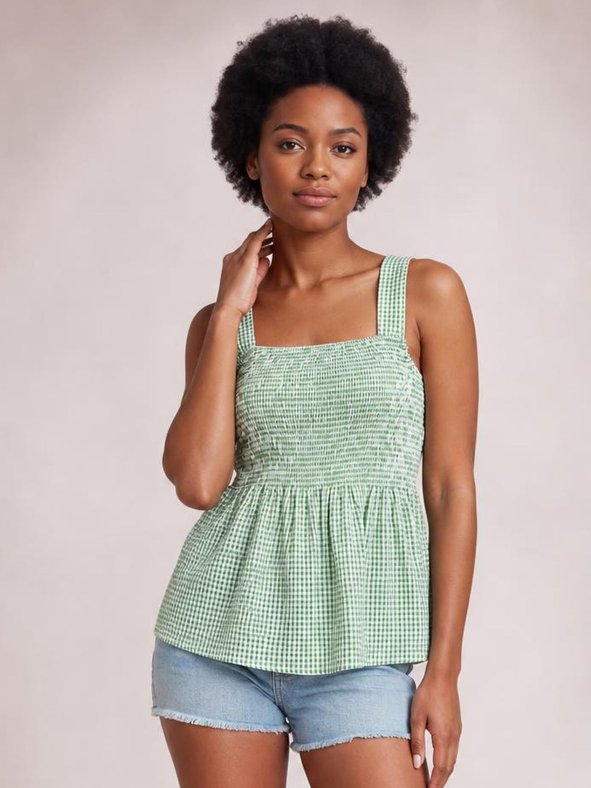 Model wearing a mint green smocked plaid tank with a square neckline, wide straps, fitted bodice, and flared peplum silhouette, touching her neck.