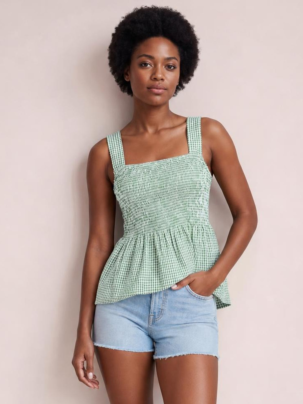 A model wears a mint green plaid tank with a square neckline, wide straps, smocked bodice, and flared peplum hem, posing with one hand in her shorts pocket.
