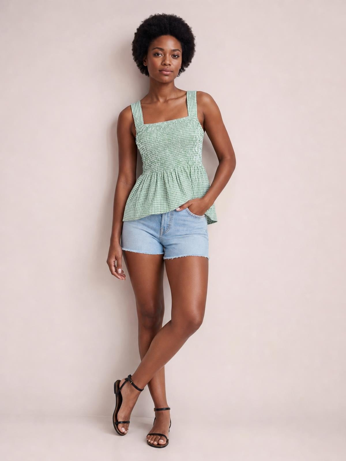 Model poses with one hand in her shorts pocket wearing a mint green plaid tank with a square neck, wide straps, smocked bodice, and flared hem.