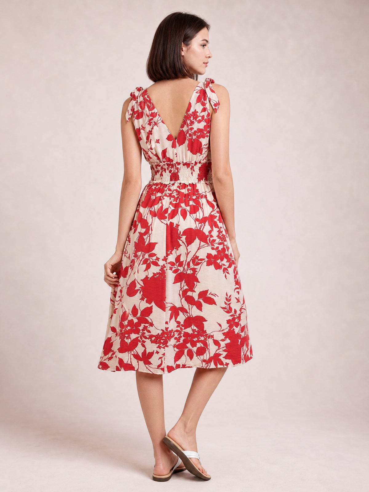 Model shows the back of a deep red floral printed midi dress with tied shoulders, a smocked waist, and a flowing A-line skirt while holding the hem.