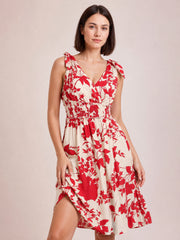 Model poses in a deep red and ivory floral printed surplice midi dress with a V-neck, tied shoulders, smocked waist, and flowing skirt.