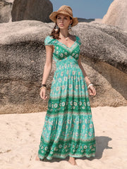 Model wears a green floral printed V-neck dress with a smocked bodice, ruched short sleeves, frill trim, and a flowing ankle-grazing tiered skirt.