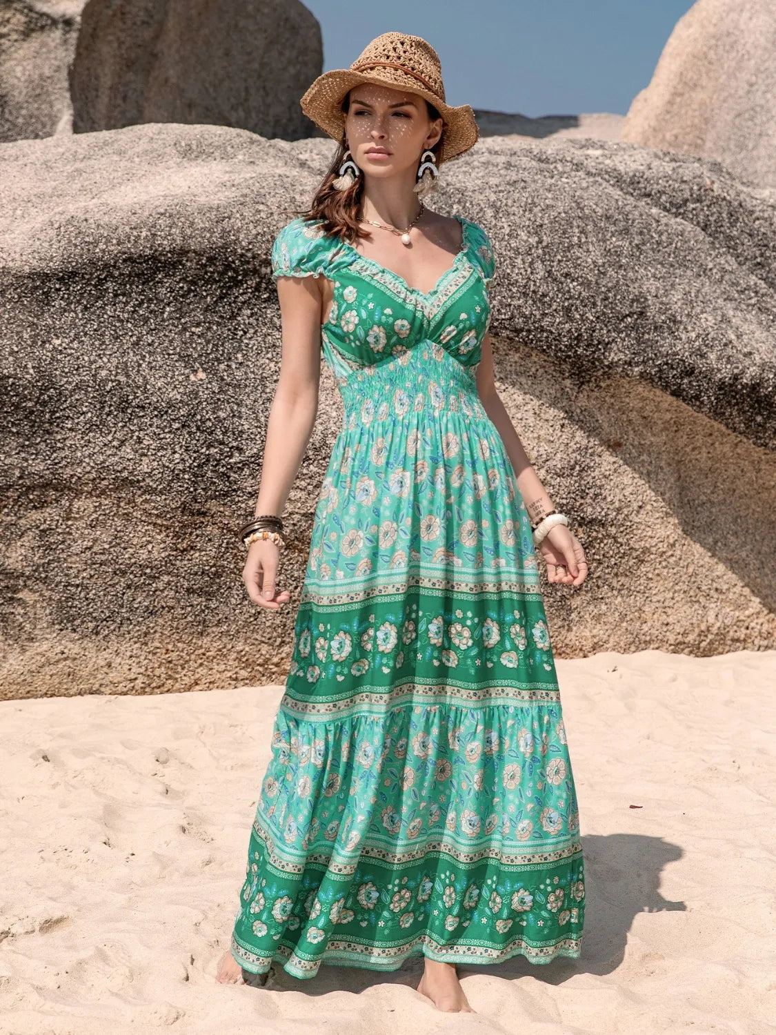Model wears a green floral printed V-neck dress with a smocked bodice, ruched short sleeves, frill trim, and a flowing ankle-grazing tiered skirt.